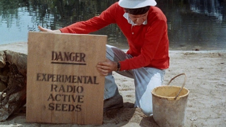 Gilligan on one knee next to a bucket and holding a sign that says 'Danger: Experimental Radioactive Seeds' in the episode 'Pass the Vegetables, Please' on Gilligan's Island