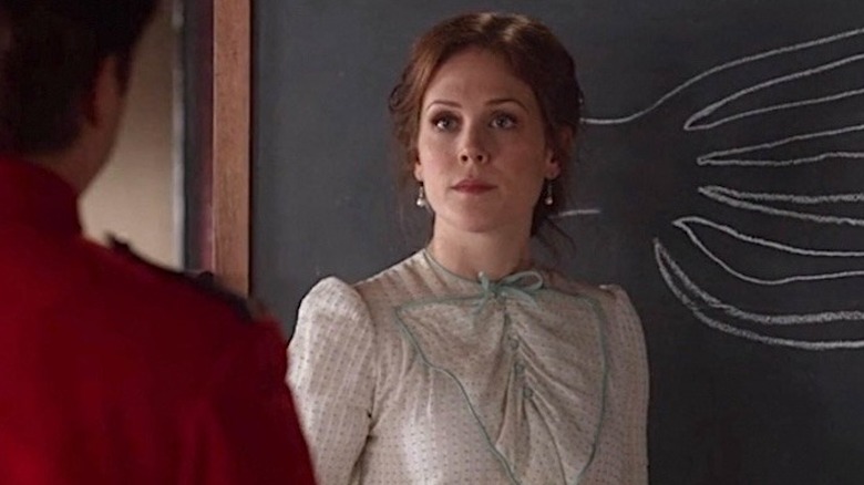 Elizabeth Thatcher in a white dress standing in front of a chalkboard facing a man in a red shirt in When Calls the Heart