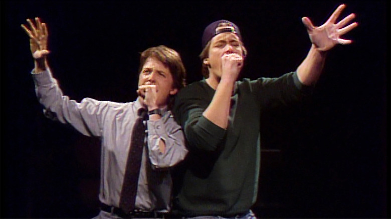 Alex P. Keaton (Michael J. Fox) and Greg (Brian McNamara) stand close together, using their hands like microphones with their other hands raised in the air as they sing against a black background