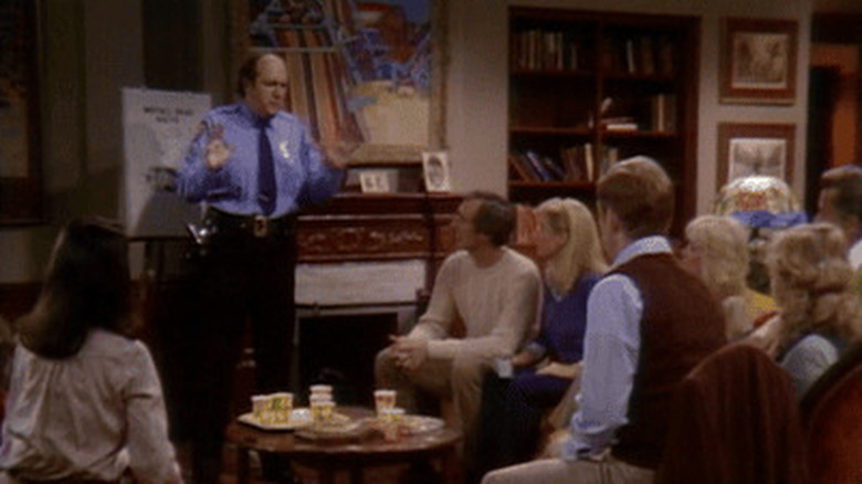 A police officer stands in the Keaton family living room in front of the fireplace, with a poster board on an easel as a group of men and women sit and listen in Family Ties