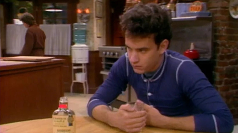Uncle Ned (Tom Hanks) sits at the kitchen table with a bottle of alcohol in front of him, wearing a grim expression, while Alex (Michael J. Fox) walks in the background in a robe in Family Ties