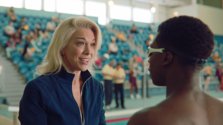 Hannah Waddingham in a tracksuit top, facing a teen in swim goggles in a pool arena with blue seating.