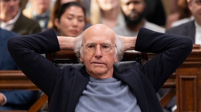Larry David sitting at a defense table in a courtroom, leaning back with his hands clasped behind his head in a scene from "Curb Your Enthusiasm."
