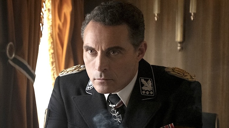 John Smith wearing a black military uniform sitting in a room in The Man in the High Castle