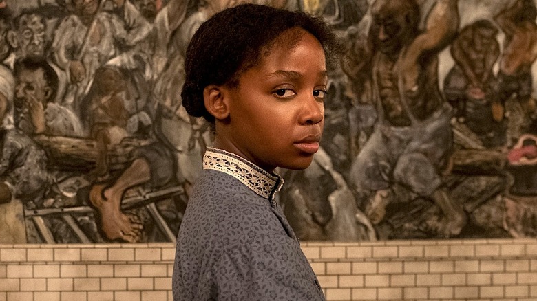 Cora in a blue dress standing in front of a wall with a mural in The Underground Railroad
