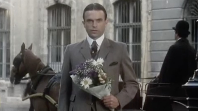 Sam Neill as Sidney Reilly with slicked back black hair wearing a gray suit holding a bunch of flowers in Reilly, Ace of Spies