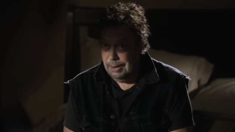 Tim Curry as Billy Flynn on Criminal Minds, sitting on a bed in the dark wearing dark clothing.