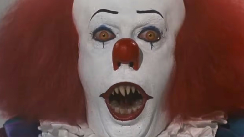 Pennywise the clown, with pale white skin, bright red hair, a bright red nose, red and yellow eyes, and pointed yellow teeth.