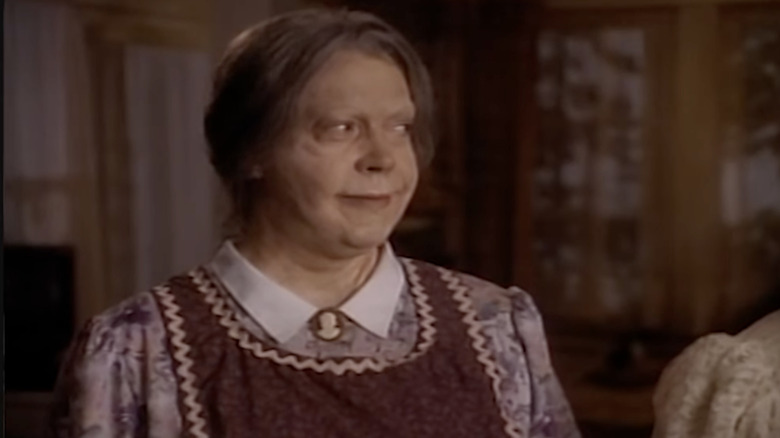 Tim Curry as an older woman wearing a purple blouse with a white collar and a dark purple pinafore.