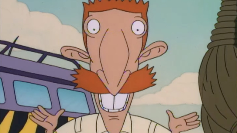 The animated Nigel Thornberry, with bright, spiky orange hair, a large mustache, large ears, and two buck teeth, is wearing a beige shirt, standing in front of a purple camping van with the light blue sky in the background.