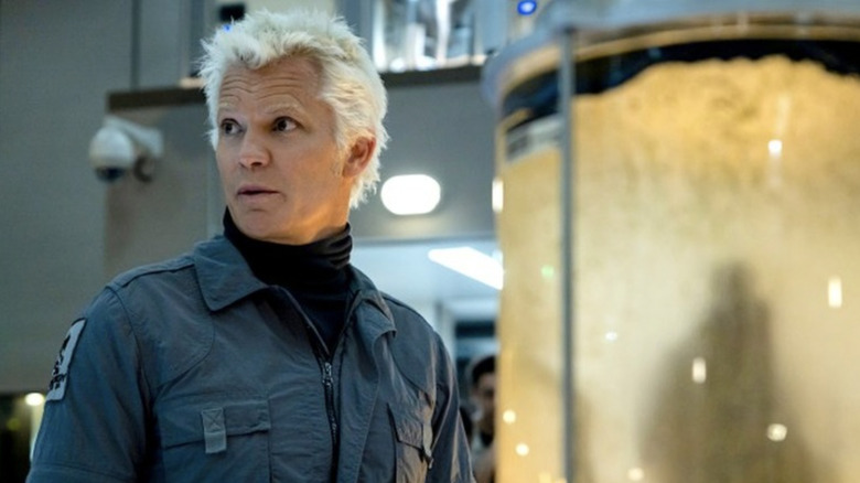 Timothy Olyphant with bleached white hair is wearing gray overalls with a black turtleneck underneath. He's looking to the left, and there is a yellow tank with something inside it behind him.