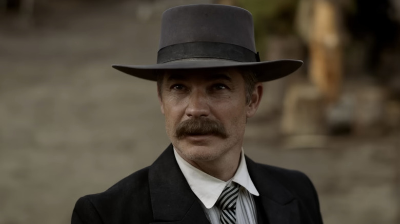 Timothy Olyphant wearing a large gray hat, a black coat, with a white shirt and black striped tie. He has a large brown mustache and is looking up at something.