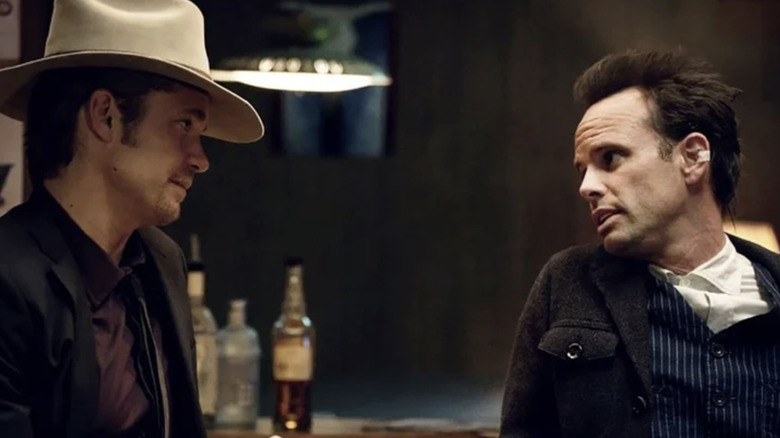Two men are sitting in a bar looking at each other. On the left the man is wearing a beige cowboy hat, a black blazer, with a maroon shirt and a dark die. On the right, the man has messy brown hair and is wearing a gray jacket, a blue striped waistcoat and a white buttoned-shirt. There is a small plaster on his ear. There are bottles of whiskey in the background.
