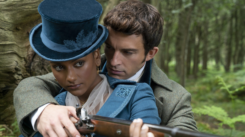 Anthony Bridgerton (Jonathan Bailey) stands closely behind Kate Sharma (Simone Ashley), helping her position and aim a hunting rifle in the woods in the series "Bridgerton."