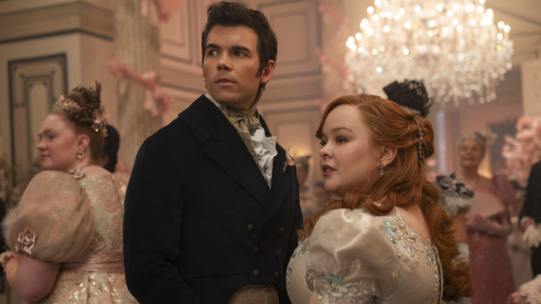 Colin Bridgerton (Luke Newton) and Penelope Featherington (Nicola Coughlan) holding hands and looking off to the side in a crowded, brightly lit ballroom.