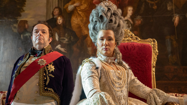 Queen Charlotte (Golda Rosheuvel) seated in an ornate, silver-and-white gown with an elaborate tall grey wig and layered pearls, alongside Brimsley (Hugh Sachs) standing formally at her side in royal uniform in the series "Bridgerton."