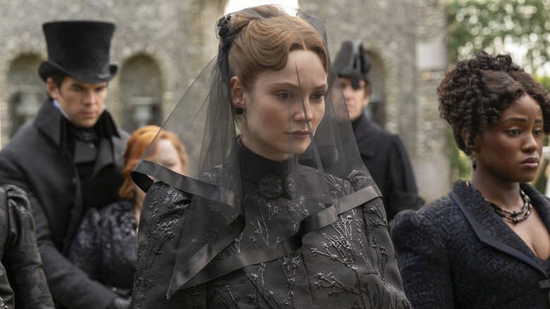 A somber Francesca Bridgerton wearing a black mourning veil and embroidered capelet, surrounded by family at a funeral in the series "Bridgerton."