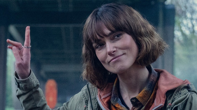 Keira Knightley waves behind her while wearing a green raincoat over a red hoodie and yellow shirt in "Black Doves."