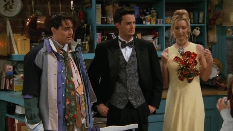 Joey wearing all of Chandler's clothes stood next to Chandler in a suit and Phoebe in a yellow dress in Friends (1996)