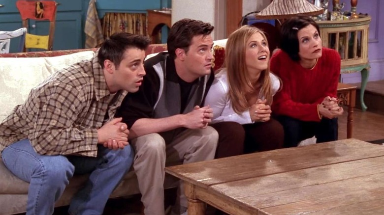 Joey, Chandler, Rachel, and Monica all sat on the apartment sofa leaning forward and looking up in Friends (1998)