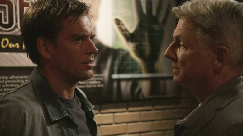 Agents DiNozzo and Gibbs talking in front of a brick wall with a poster on "NCIS"