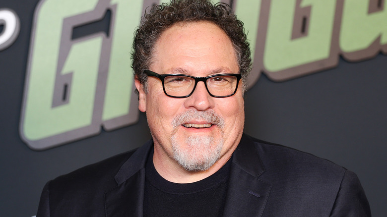 Jon Favreau in a black suit and glasses in front of a black and green background