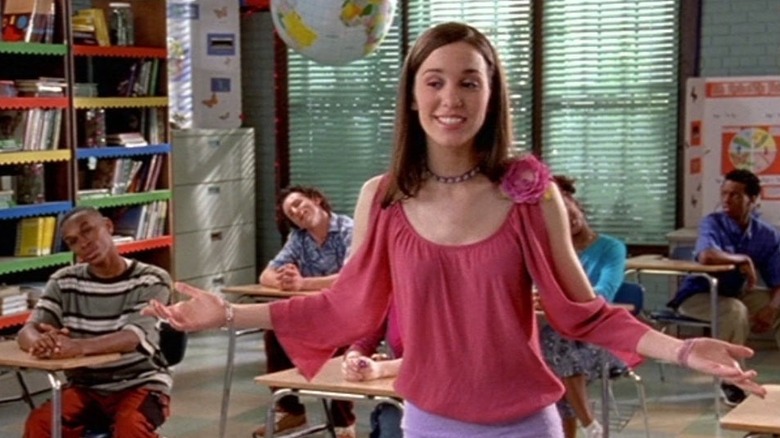 Ren (Christy Carlson Romano) stands with her arms outstretched, wearing a pink shirt with a flower, while students sit at their desks with their hands folded.