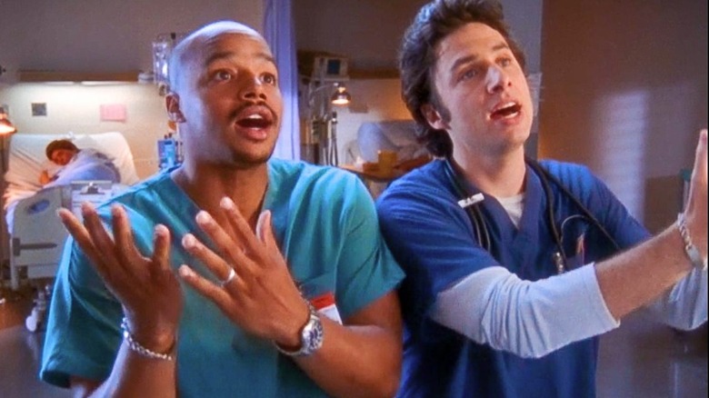 Turk (Donald Faison) and J.D. (Zach Braff) stand side by side in their scrubs, both with their hands raised and mouths open mid-song, with the hospital in the background