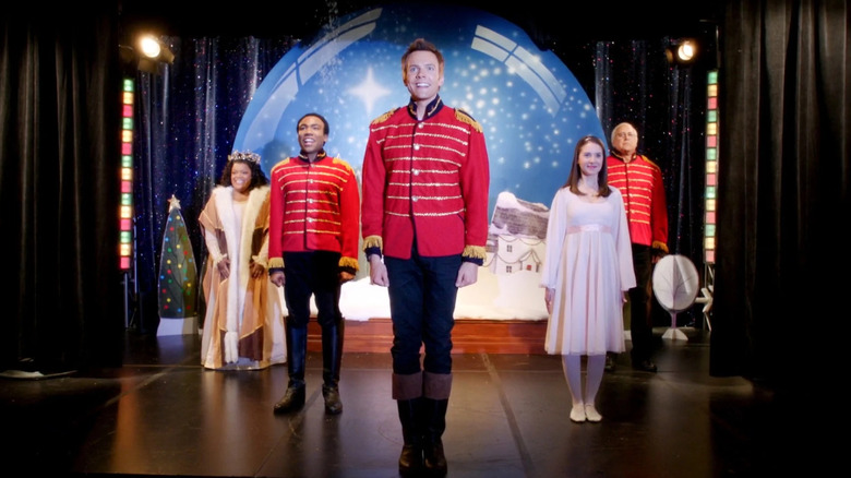 The Community ensemble stands onstage in Christmas-themed outfits, with a winter snow globe backdrop behind them.