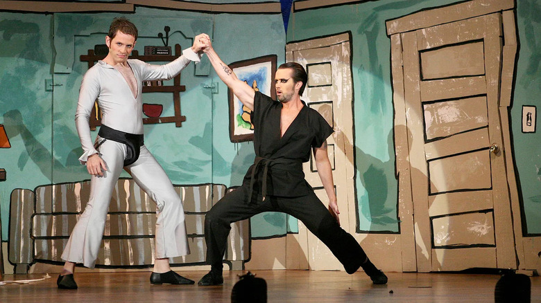 Dennis (Glenn Howerton), wearing a silver bodysuit, and Rob McElhenney (Mac), performing as the Nightman, hold hands and stand in a wide-legged stance against a crayon-style living room set