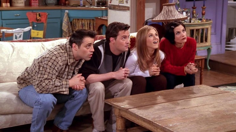 Joey Tribbiani, Chandler Bing, Rachel Green, and Monica Geller sitting on the couch and looking up at someone eagerly in Friends