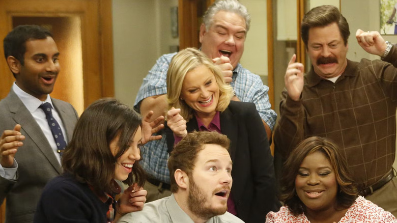 Tom Haverford, April Ludgate, Andy Dwyer, Leslie Knope, Garry Gergich, Ron Swanson, and Donna Meagle looking down at something on a table in Parks and Recreation