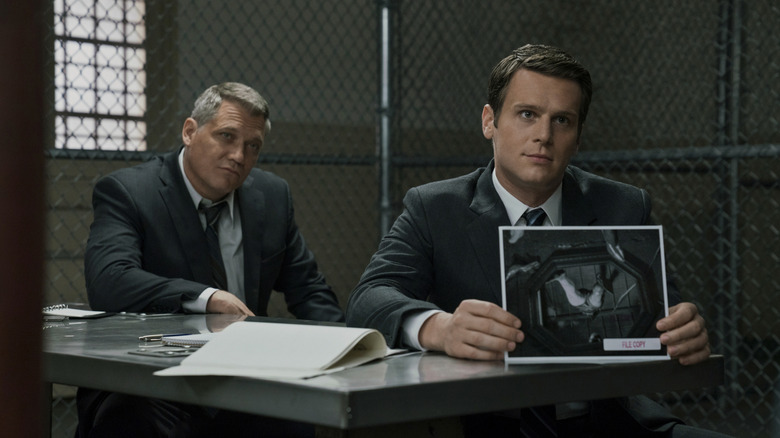 Two men in matching dark suits sit at a metal tale in prison. On the left, the older man has short graying hair and is clean shaven. On the right, the younger man has neat brown hair and is holding up a black-and-white crime scene photo.