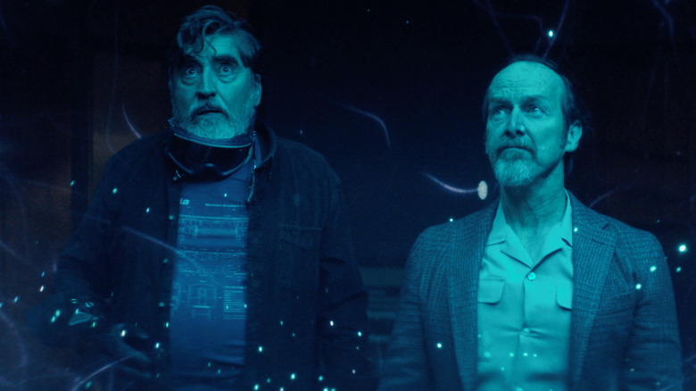 Two older men are illuminated by glowing blue light. On the left, the older man has messy hair and a fringe. He's wearing dark shirt over the top of a T-shirt. There are a pair of goggles around his neck. On the right, the man has swept back hair with a gray blazer over the top of a beige cuban collared shirt.