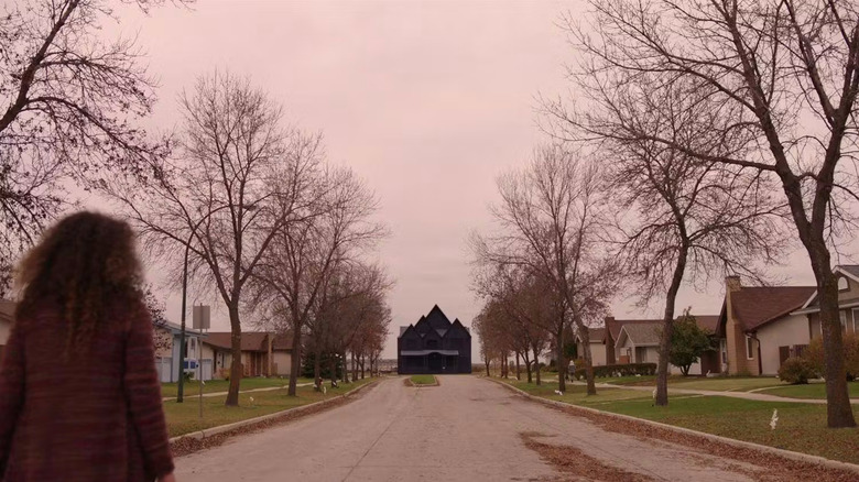 A girl walking down a street lined with houses and an ominous black house is at the foot of the street in Channel Zero: No-End House