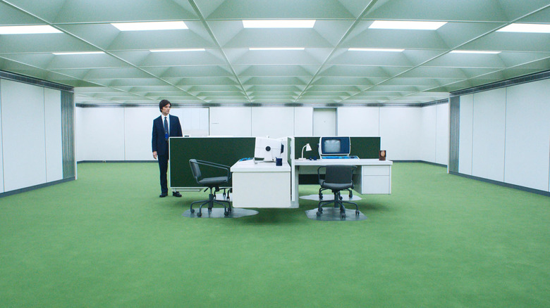 Mark Scout standing next to desks in a Lumon office space room with green flooring in Severance