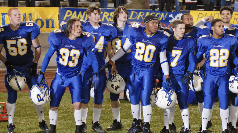 Group of football players in blue uniforms with white and yellow numbers, holding helmets.