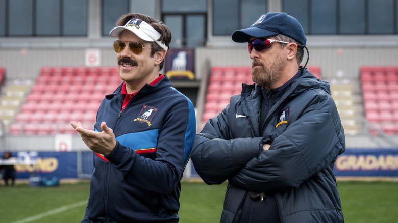Jason Sudeikis as Ted Lasso and Brendan Hunt as Coach Beard in tracksuits and caps.