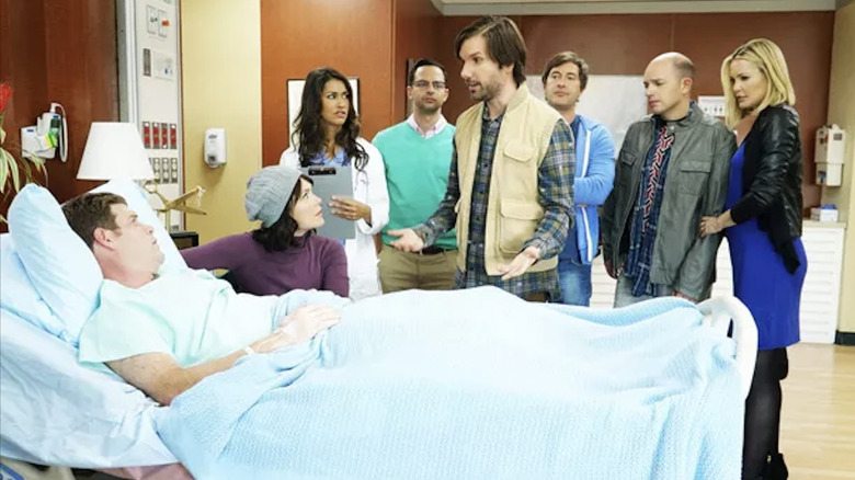 A group of friends all stand around a man lying in a hospital bed.