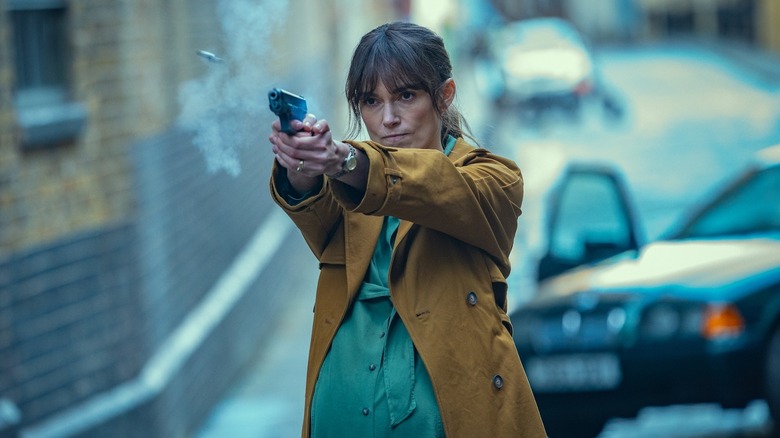 Keira Knightley as Helen Webb, she has brown hair and is wearing a yellow coat with a green blouse underneath, she's pregnant and firing a pistol in the street.