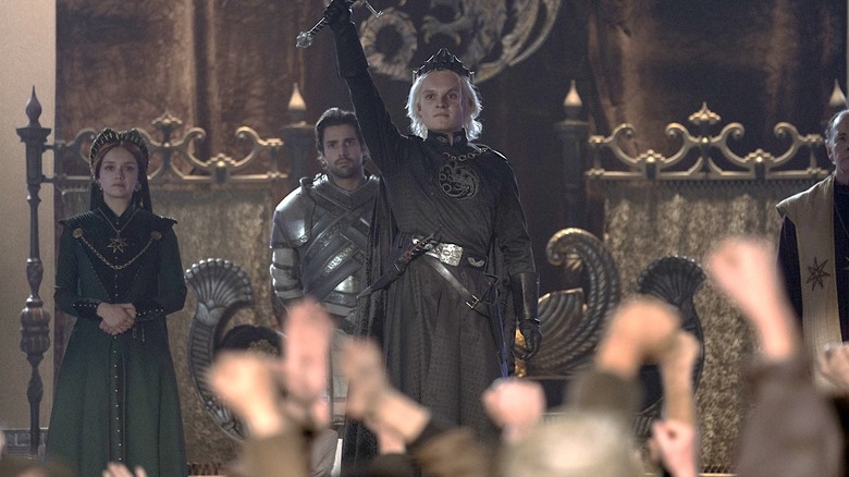 Aegon II holding up a sword in front of a crowd with Alicent Hightower and Criston Cole in the background