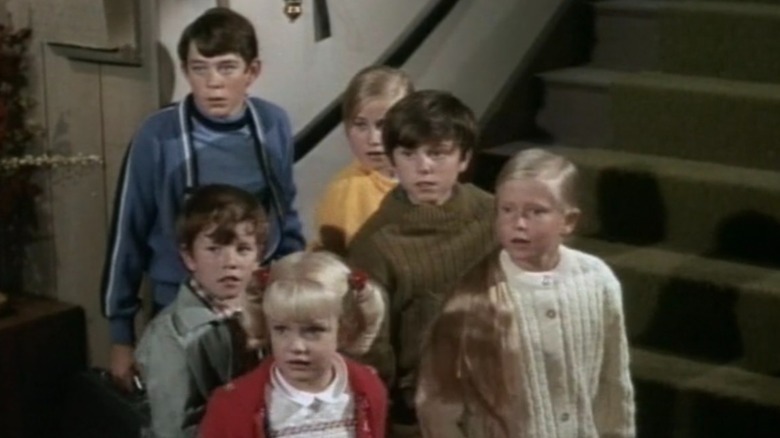 The Brady Bunch standing in front of a staircase looking back at something with surprise.
