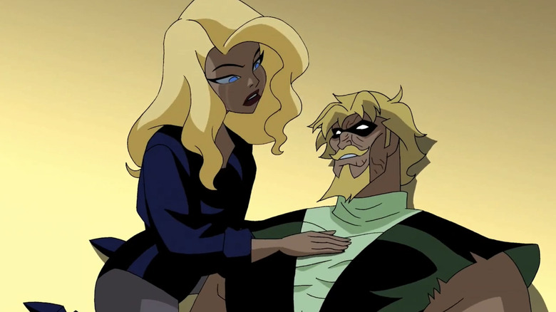 Two blond superheroes. On the left, Black Canary is wearing a black leather jacket over the top of a black leotard and tights. She doesn't have a mask on. She's sitting with her hand on Green Arrow's chest, who is laying down on the floor next to her. He has a blond goatee and a black mask with white lenses, and is wearing a two-toned green costume that is ripped following a fight.
