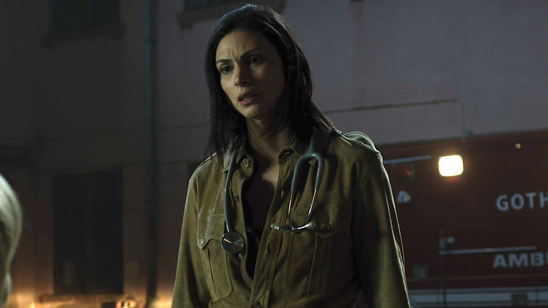 A woman with black hair and a green shirt. She has a stethoscope around her neck. She is standing in a warehouse, and there is a red and white ambulance behind her.