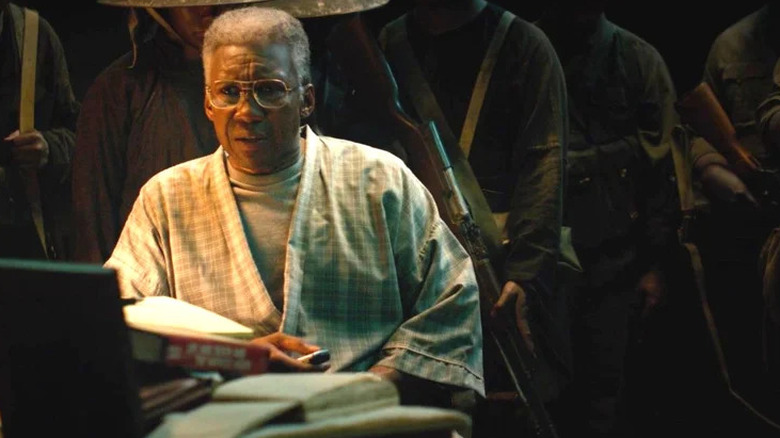 An older Wayne Hays (Mahershala Ali) sits on a desk piled with books and legal pads, wearing a robe, with Viet Cong soldiers behind him in dark lighting in True Detective