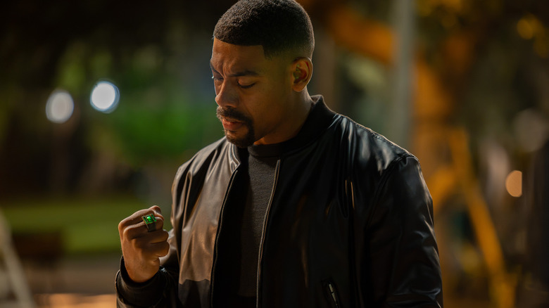 Aaron Pierre as John Stewart with short black hair and a goatee wearing a black leather jacket and looking at a green ring on his finger in Lanterns.