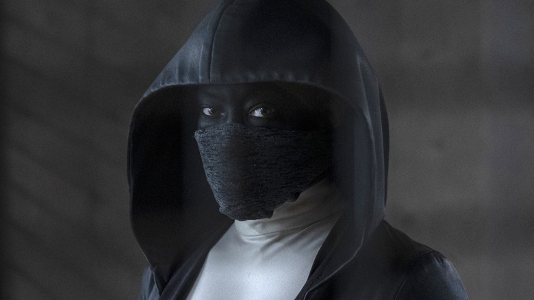 Regina King as Sister Night wearing a black hood with a face covering and her face painted black. She's wearing a white top underneath the black robe.