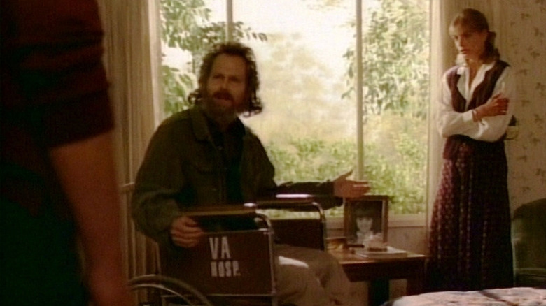 Jeff in wheelchair in bedroom with Denise standing nearby with arms folded