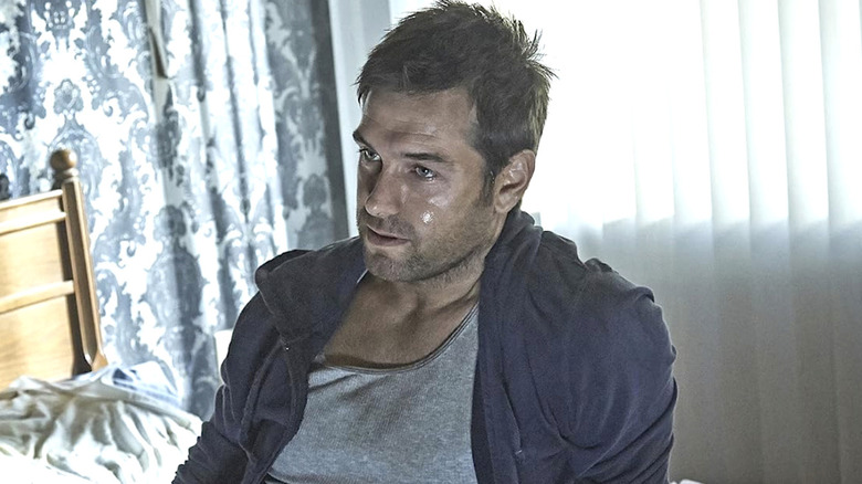 Lucas hood wearing a grey shirt and blue jacket sitting on a bed with sweat on his face in "Banshee"