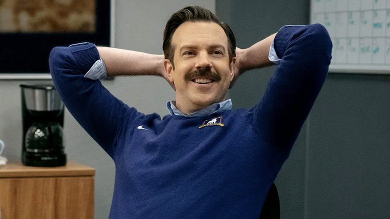 Ted Lasso lounges in an office chair with his hands behind his head on the Apple TV series.